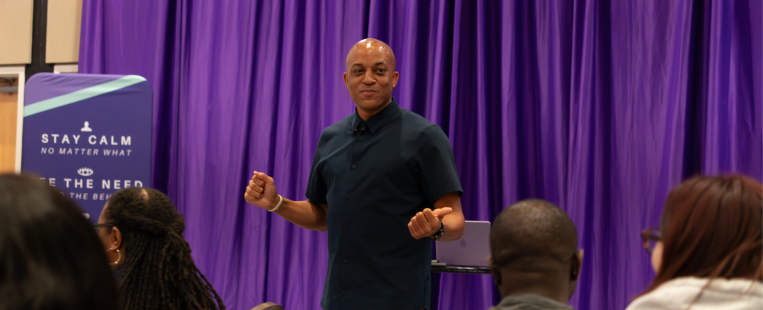 A person stands confidently in front of an audience, gesturing with hands, against a backdrop of purple curtains. A banner to the left reads "STAY CALM NO MATTER WHAT." Attendees face the speaker, attentively listening.