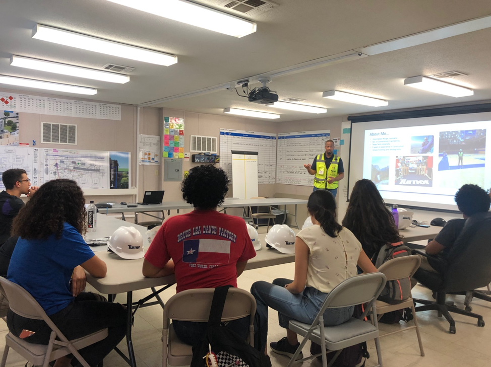 A person dressed in a yellow safety jacket giving a presentation to a group of students with safety helmets on their desks