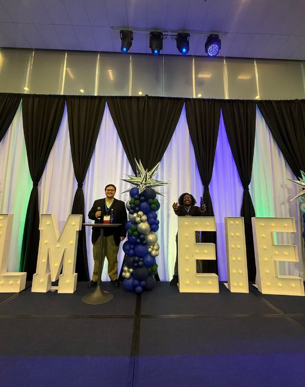 Two individuals stand on a stage decorated with large illuminated letters spelling "IELM." The backdrop features dark curtains with a star-topped balloon arrangement in the center. The setting is festive, with bright lighting and a sense of celebration.