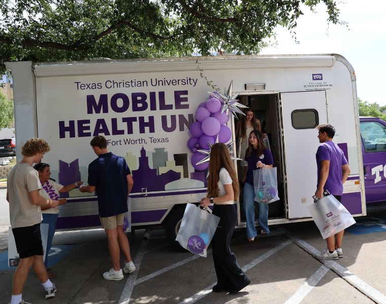 A group of people stand outside the Texas Christian University Mobile Health Unit in Fort Worth, Texas. They appear to be engaged in conversation and carrying bags. The vehicle is decorated with purple balloons, and a few individuals are stepping in and out of the unit.