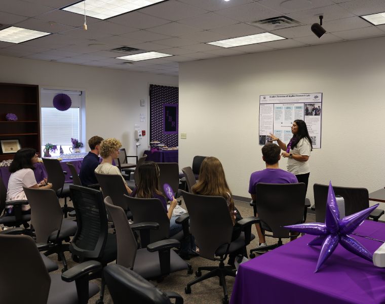 A person stands at the front of a room, presenting to a seated audience. The room has several chairs, some occupied. Purple decorations are present, such as balloons and streamers.