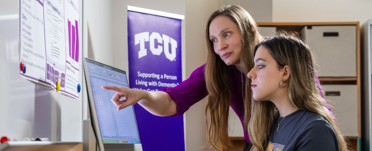 Two people are working at a computer in an office. A screen displays a data sheet. A TCU banner in the background reads 'Supporting a Person Living with Dementia? We’ve Got Tools to Help!'
