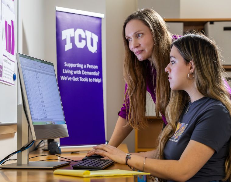Two people are working at a computer in an office. A screen displays a data sheet. A TCU banner in the background reads 'Supporting a Person Living with Dementia? We’ve Got Tools to Help!'