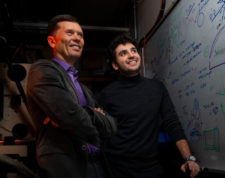 Two individuals are standing in front of a whiteboard covered with complex mathematical formulas and diagrams. They appear engaged and thoughtful, in a room with various technical equipment.