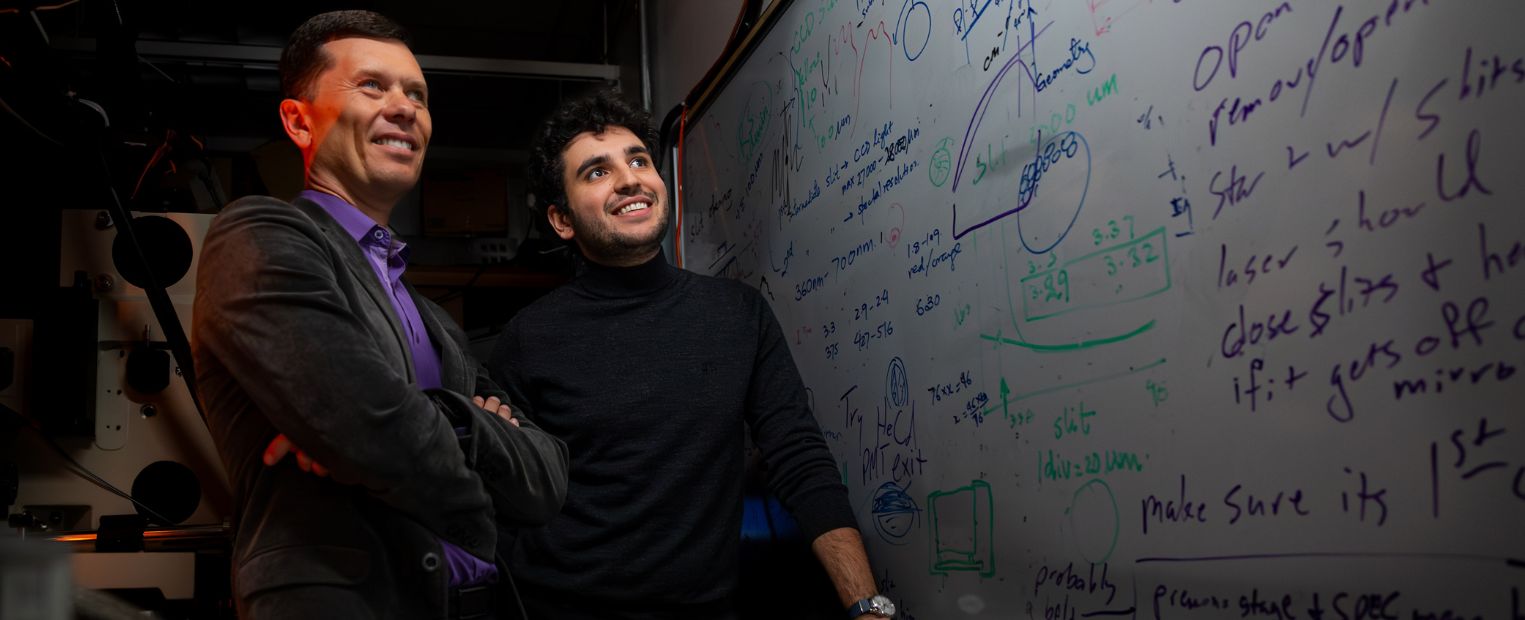 Two individuals are standing in front of a whiteboard covered with complex mathematical formulas and diagrams. They appear engaged and thoughtful, in a room with various technical equipment.