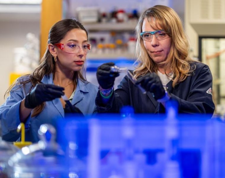 Two people in a laboratory setting, wearing safety goggles and lab coats, are handling scientific equipment. Shelves with various bottles and tools are visible in the background.