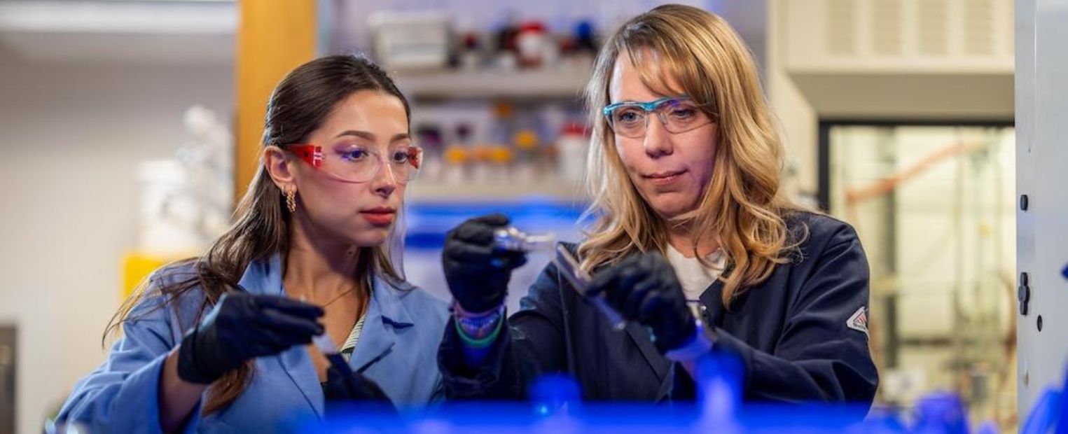 Two people in a laboratory setting, wearing safety goggles and lab coats, are handling scientific equipment. Shelves with various bottles and tools are visible in the background.