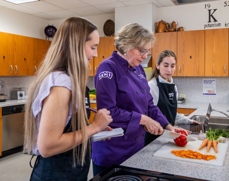 A culinary instructor from TCU demonstrates cutting a red bell pepper in a kitchen. Two students observe and take notes. Fresh carrots are also on the counter. Kitchen cabinets and a periodic table element sign on the wall are visible in the background.