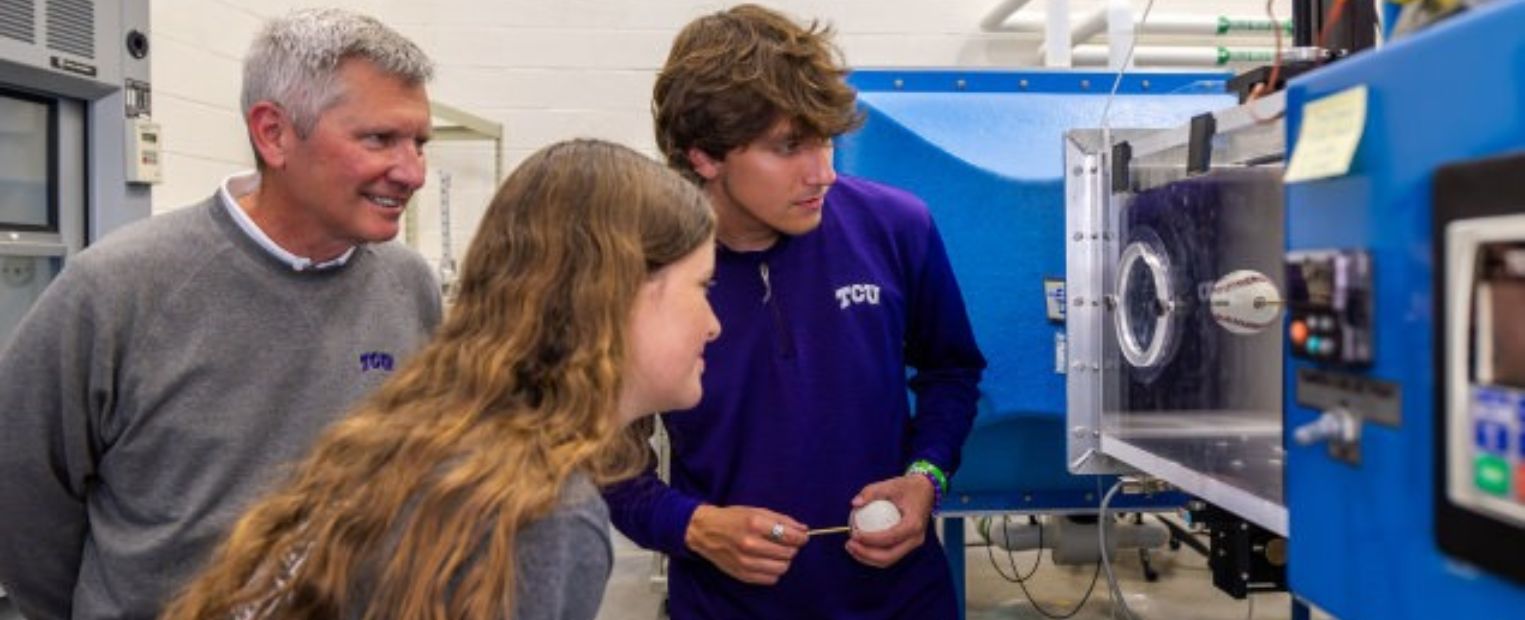 Three people are observing a scientific experiment involving air flow around a baseball in a laboratory setting. One of them is holding a baseball while looking at data on a machine. They are wearing casual clothing with "TCU" logos. The room is equipped with various lab equipment and machinery.