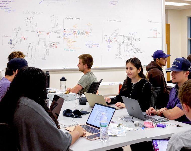 A group of students is seated around a table, each with a laptop. Behind them is a large whiteboard covered in detailed diagrams and drawings. Some students are focused on their screens while others are engaged in conversation. The setting appears to be a classroom or study area.