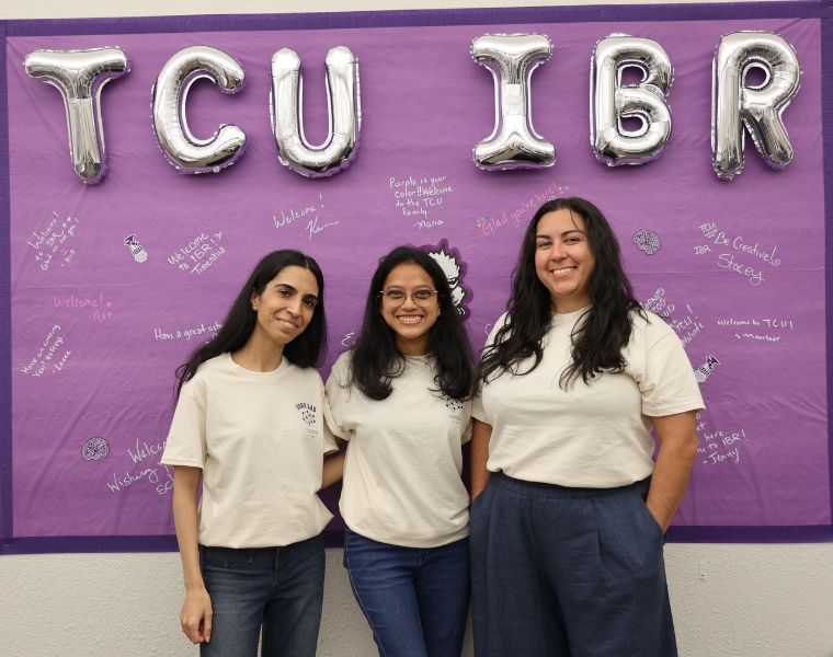 Three students stand in front of a purple banner, with balloons spelling out "TCU IBR"