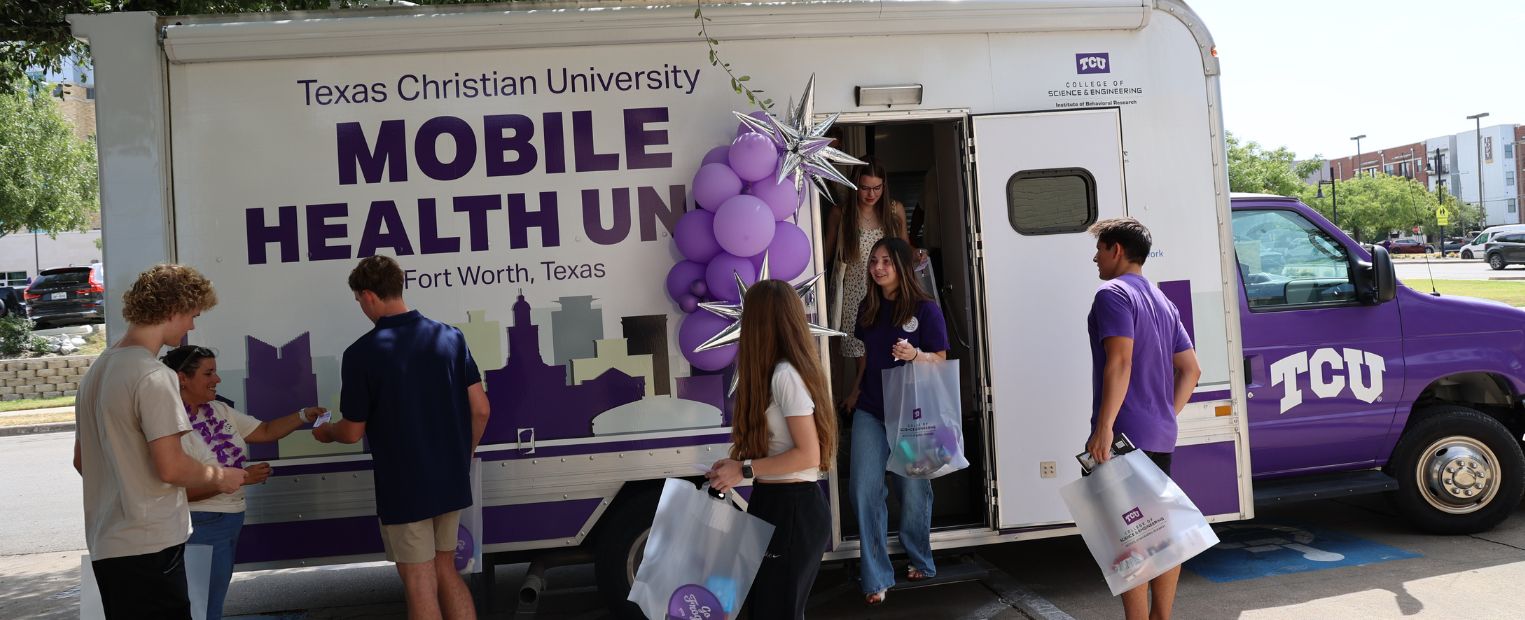 A group of people stand outside the Texas Christian University Mobile Health Unit in Fort Worth, Texas. They appear to be engaged in conversation and carrying bags. The vehicle is decorated with purple balloons, and a few individuals are stepping in and out of the unit.