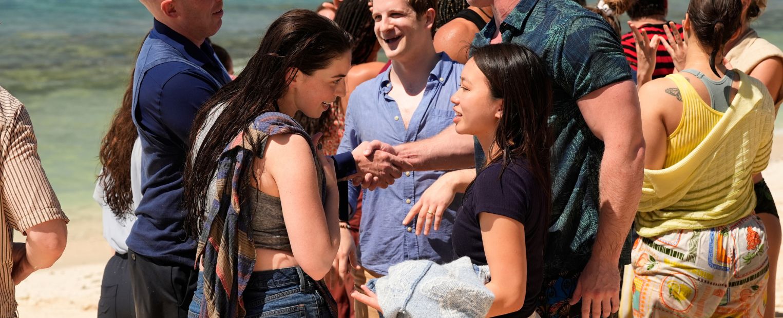 A group of people are gathered and smiling on a beach with clear blue water and distant islands in the background. They appear to be enjoying each other's company, with several involved in conversation. The scene is bright and sunny, suggesting a warm and pleasant day.