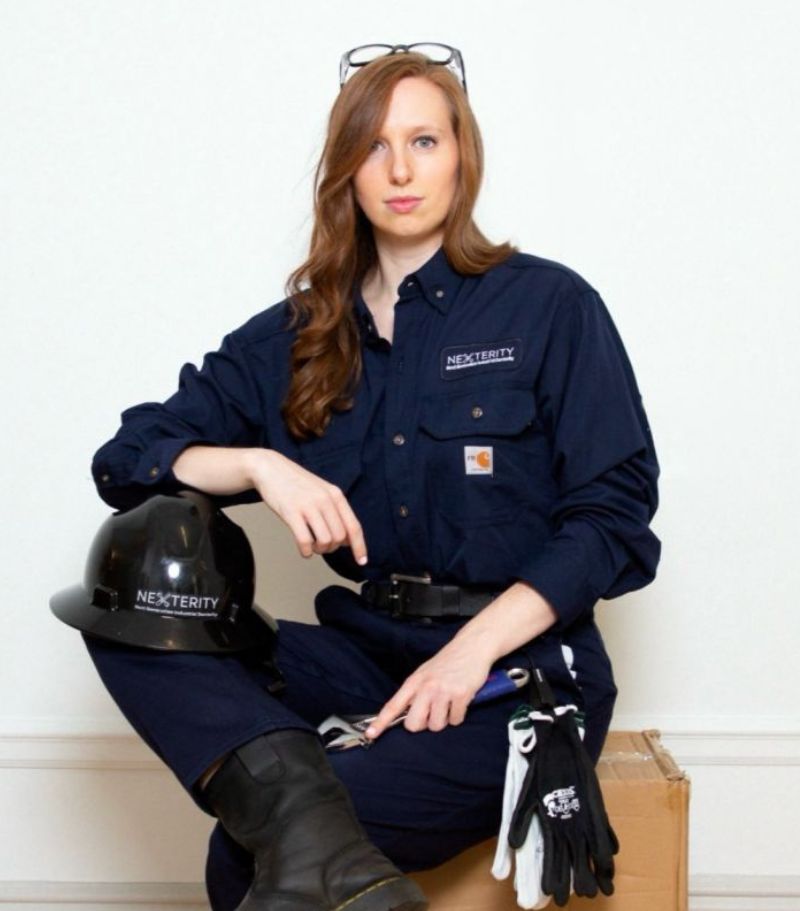 An individual wearing a dark blue work uniform with "Nexterity" logo, seated on a cardboard box. A hard hat with the Nexterity logo is placed nearby, along with gloves and safety glasses.